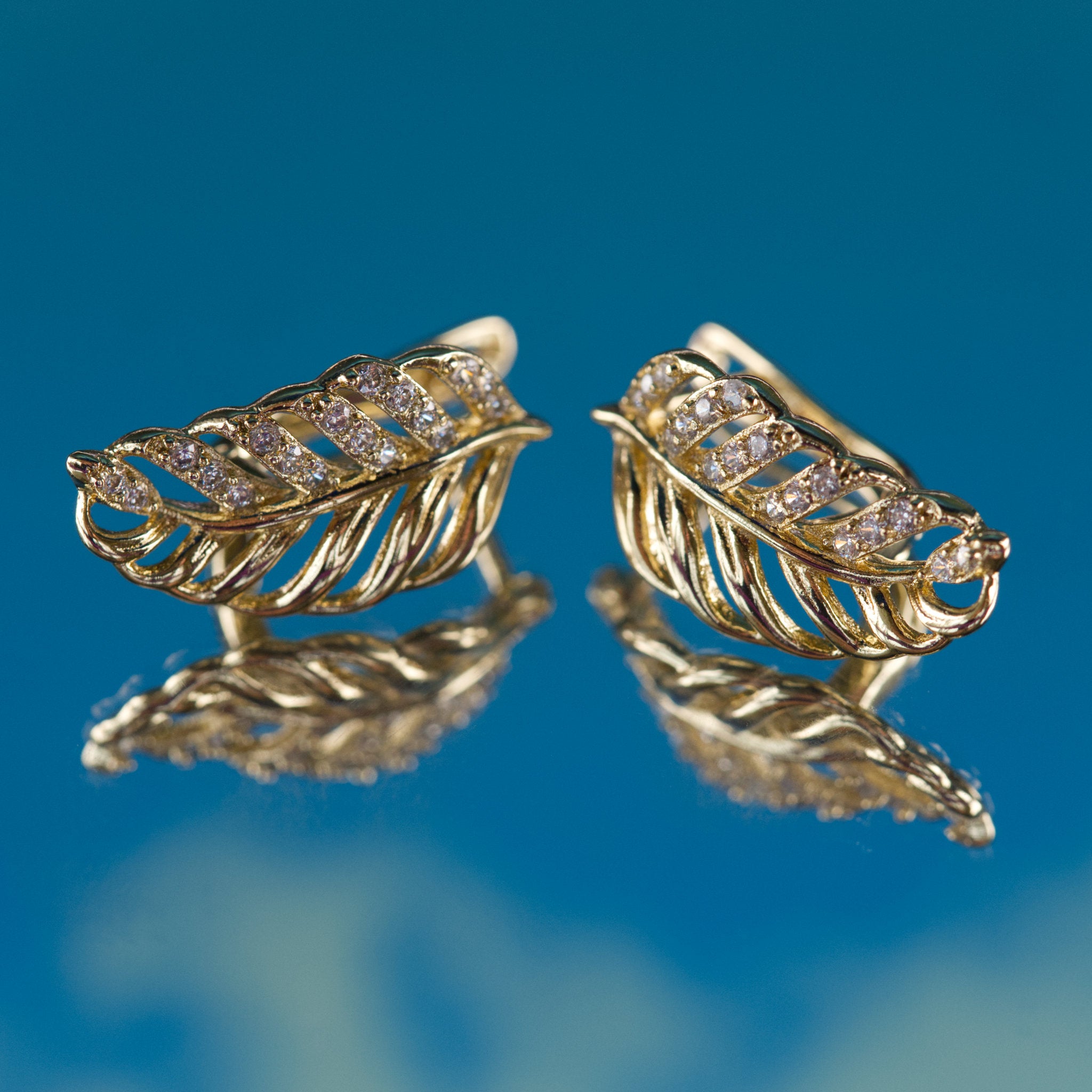Gold feather-shaped earrings with diamond accents on a blue background