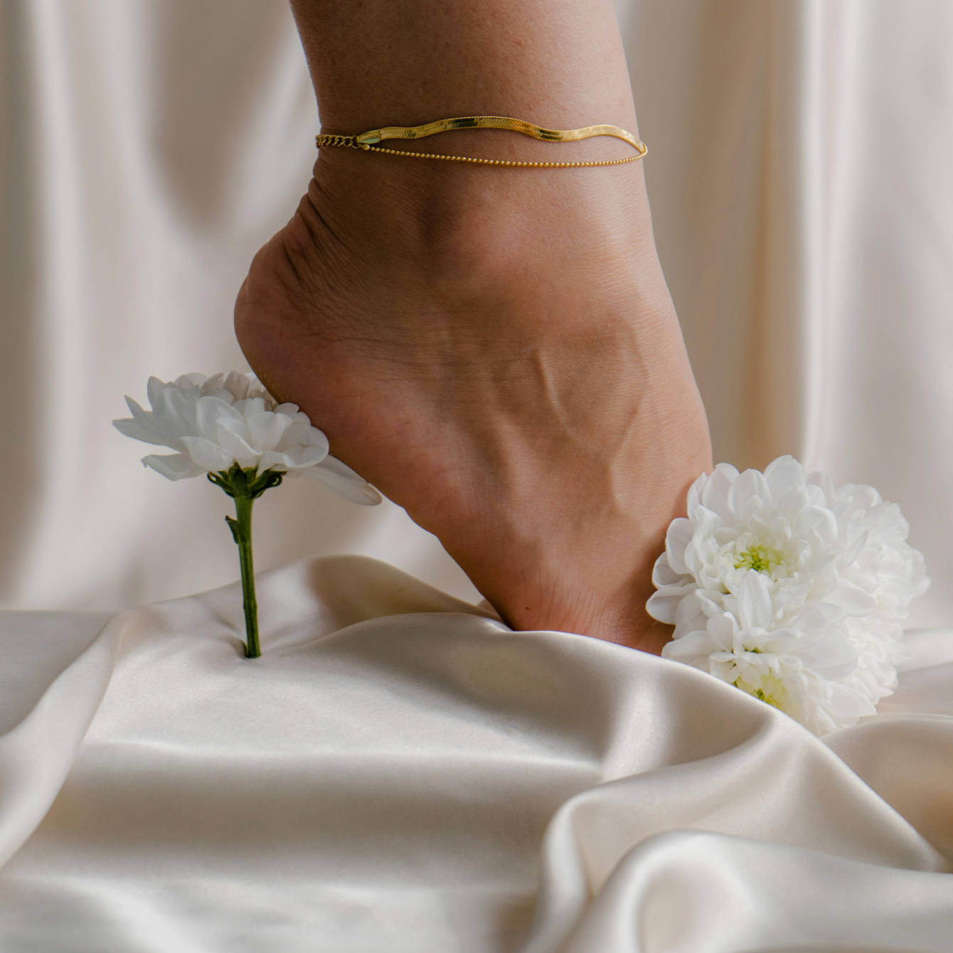 Gold anklet on a foot with white flowers on a light fabric background